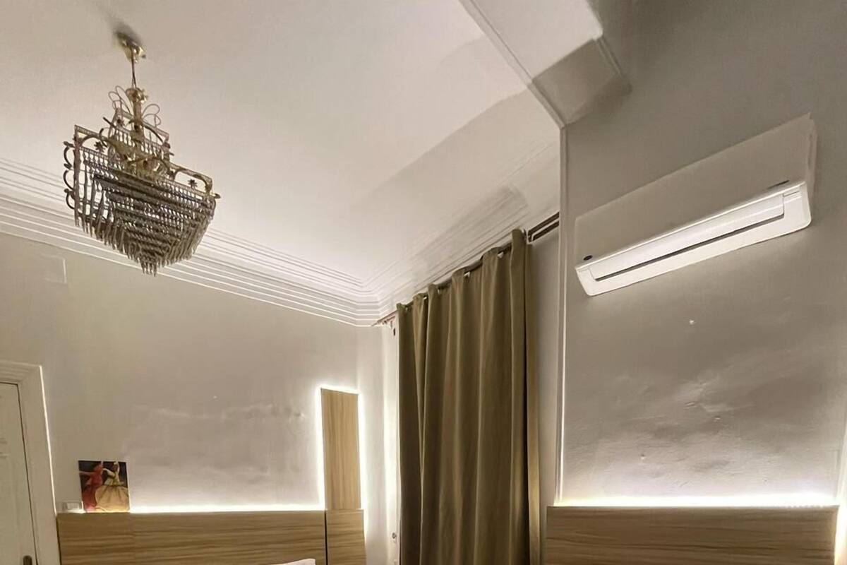A hotel room with a large bed, a smaller bed, a nightstand, a lamp, a chandelier, and a window with curtains.
