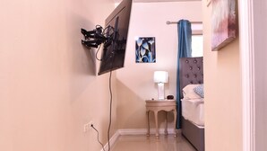 Ein Hotelzimmer mit Flachbildfernseher an der Wand, einem Nachttisch mit Lampe, einem Bett mit gestepptem Kopfteil und einem Bild an der Wand.