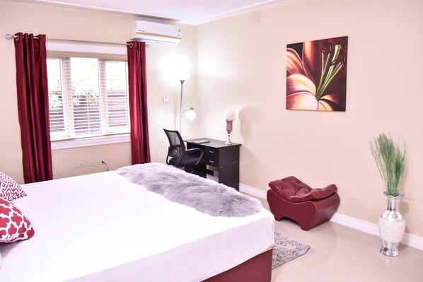 A bedroom with a bed, a desk, a chair, a red chair, a vase with plants, and a painting on the wall.