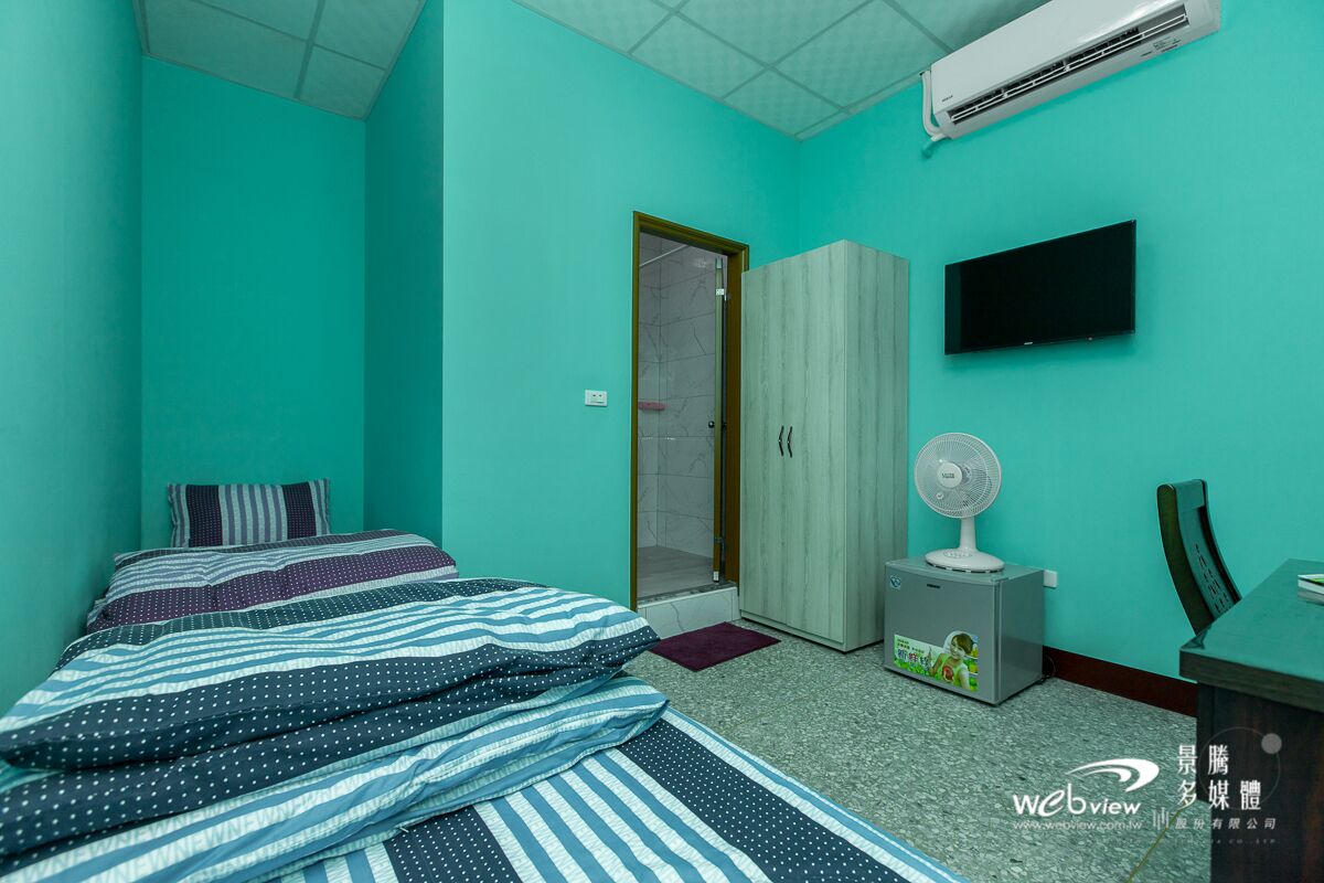 Quarto com paredes azul-esverdeadas, cama com roupa de cama listrada, guarda-roupa, televisão, ventoinha e ar condicionado.