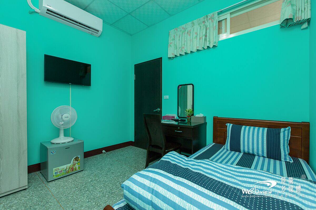 A room with teal walls, a bed with striped bedding, a desk with a chair, a TV mounted on the wall, and an air conditioner.