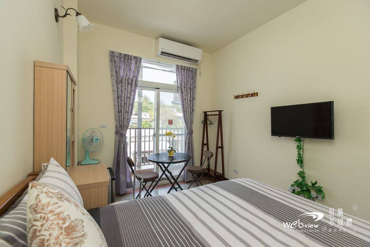 A bedroom with a bed, a TV mounted on the wall, a small table with chairs, and an air conditioning unit.