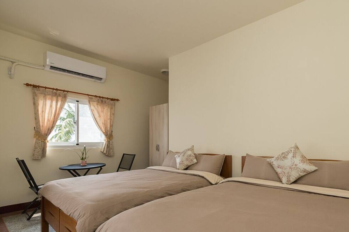 A hotel room with two beds, a desk, an air conditioner, and a window with curtains.