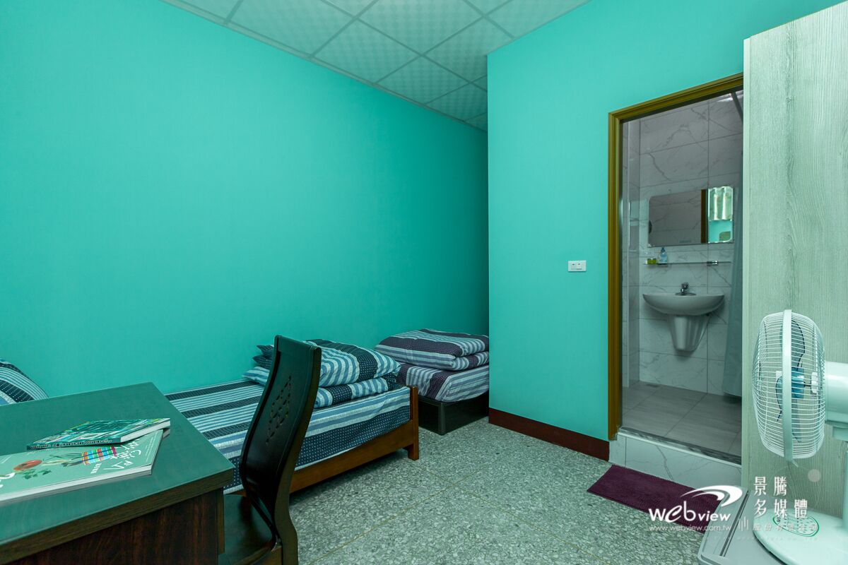Un alojamiento pequeño y de una sola habitación con paredes color turquesa, un escritorio de madera, una silla, dos camas, un lavabo pequeño y un ventilador de pie.