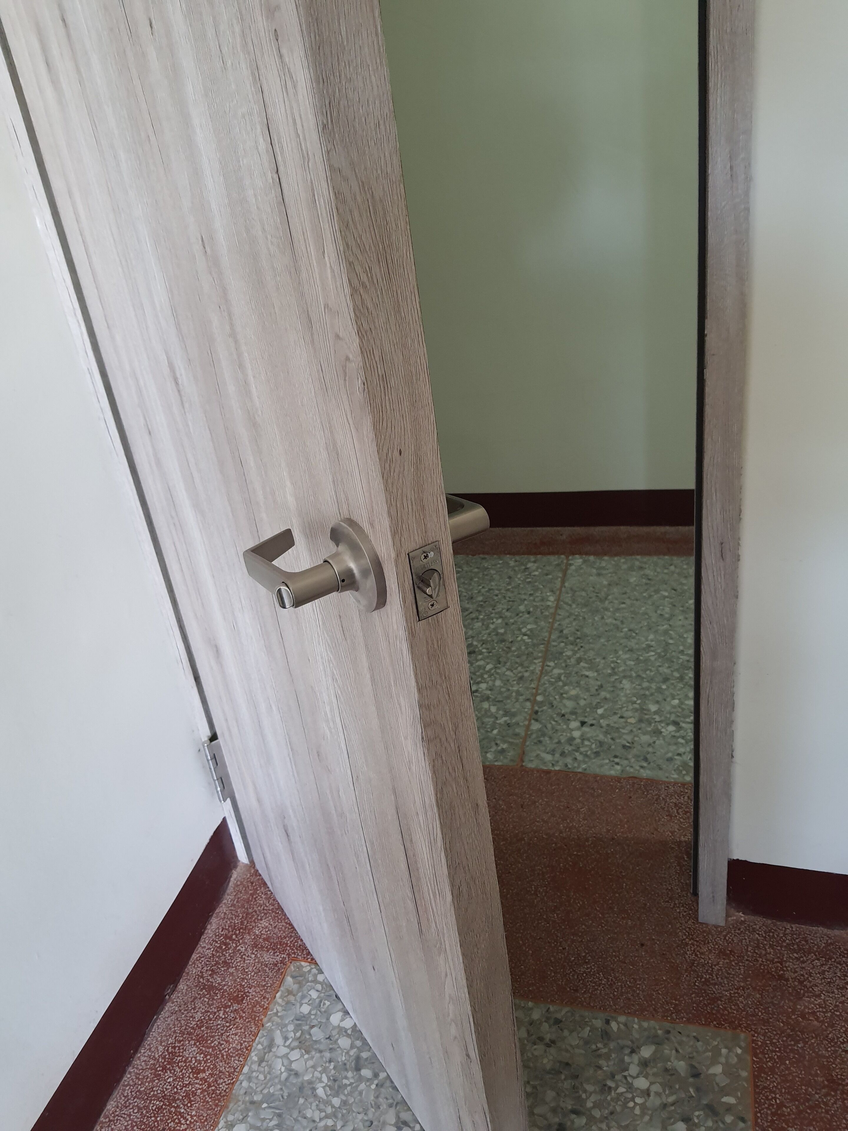 Una puerta de madera con manija metálica y cerradura, que da a una habitación con piso de baldosas y pared verde.
