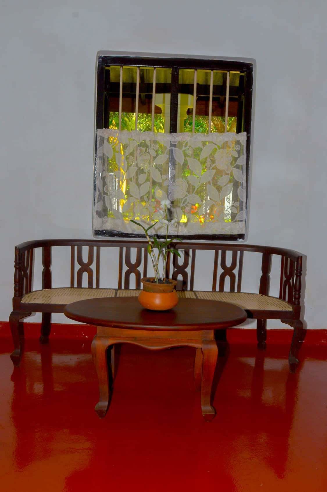 A wooden bench with a woven seat, a round wooden table with a potted plant, and a window with decorative bars and a lace curtain.