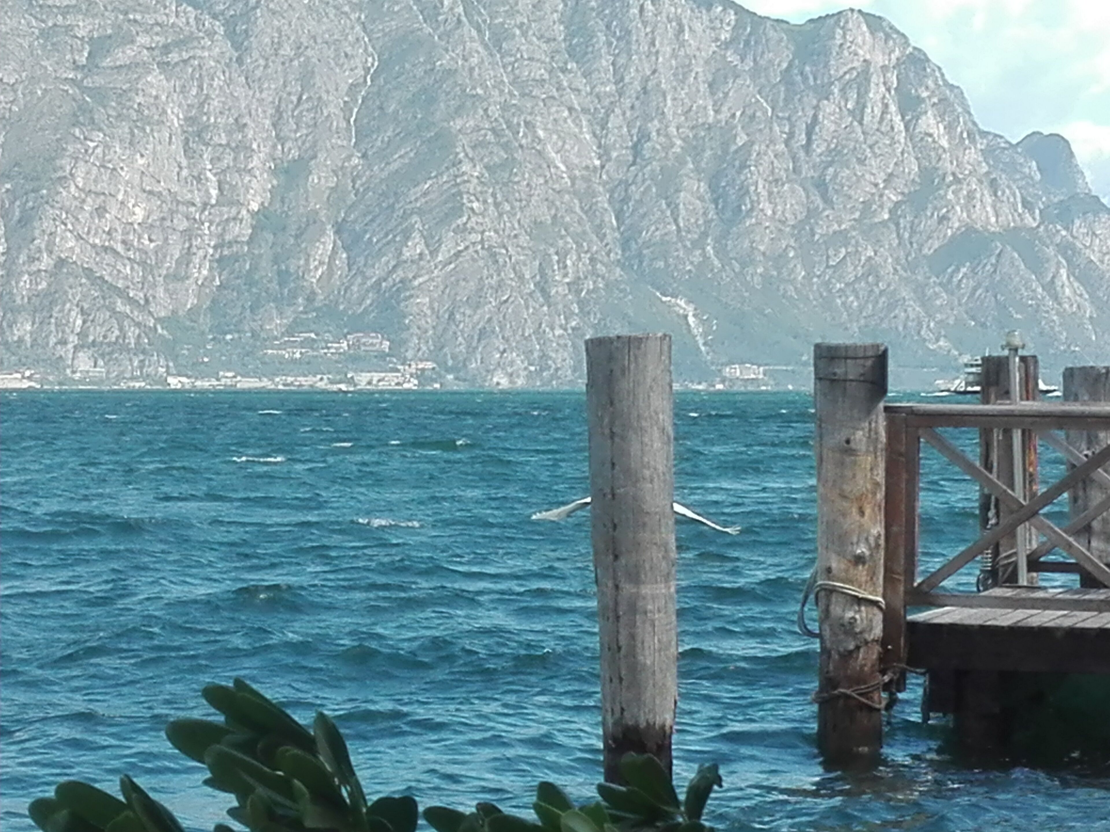 Escena costera con un muelle de madera que se adentra en el mar, rodeado de acantilados rocosos y una costa lejana con edificios.