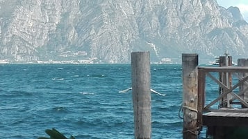 Escena costera con un muelle de madera que se adentra en el mar, rodeado de acantilados rocosos y una costa lejana con edificios.
