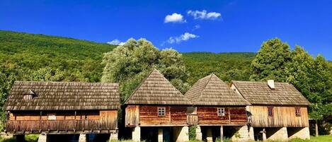 A row of rustic wooden houses with thatched roofs, situated by a river, under a clear blue sky.