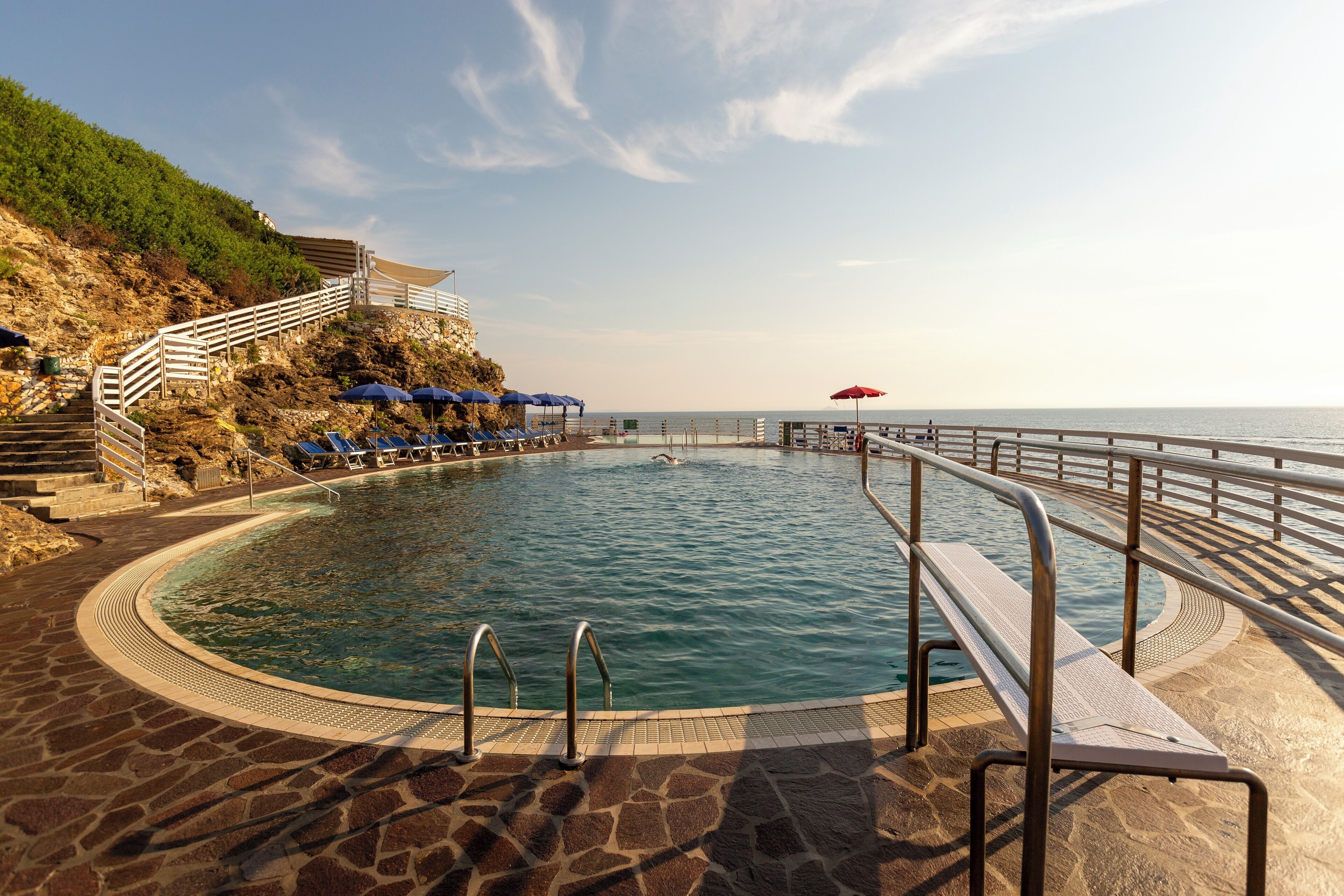 Zona de piscina con solárium de piedra, barandilla metálica y vista al mar.