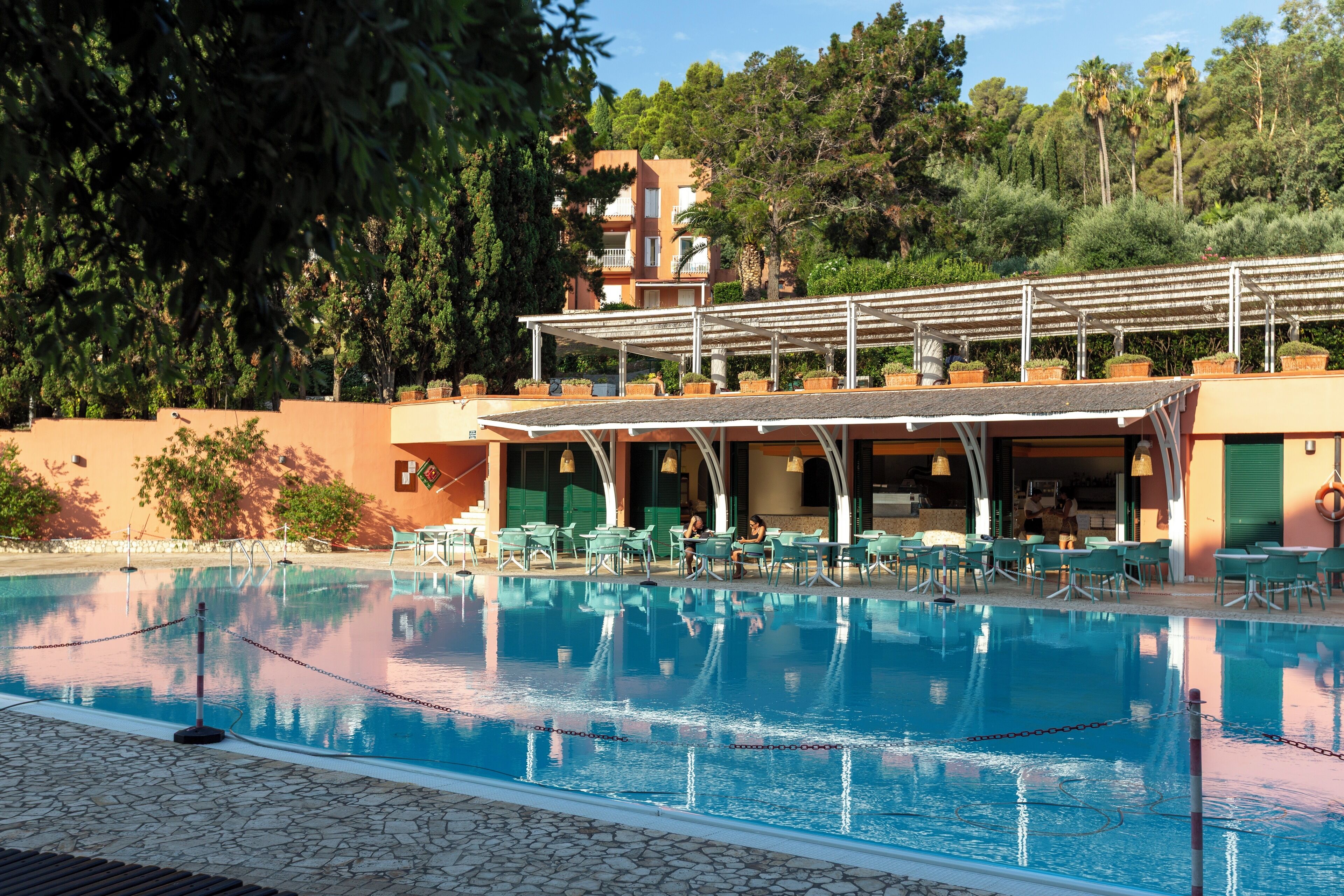 Une piscine entourée de chaises longues et de tables, elle-même bordée de verdure et d’un bâtiment doté de balcons.