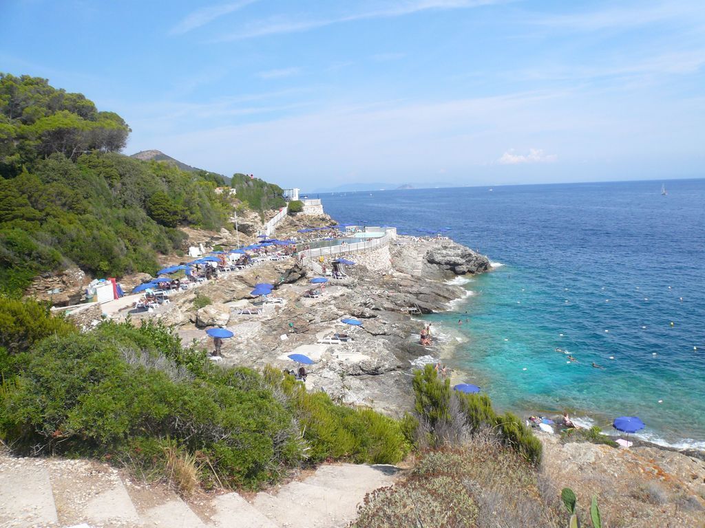Une zone côtière caractérisée par un terrain rocheux, des eaux d’un bleu limpide et une rangée de parasols le long de la plage.