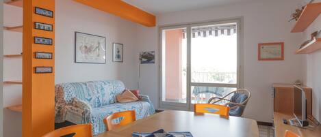 A dining area with a table, chairs, and a sofa. There are framed pictures on the wall and a window with a view.
