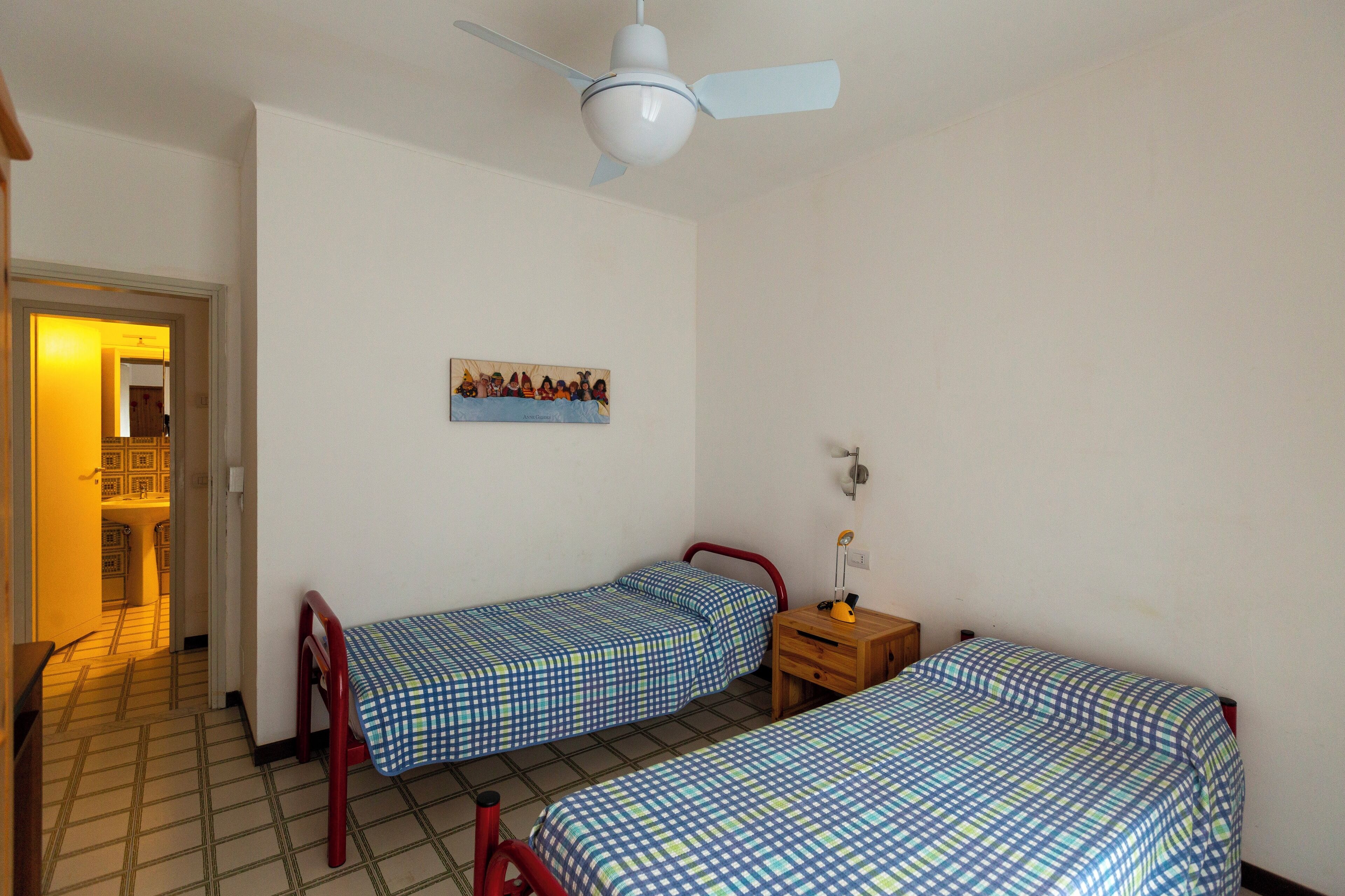 Une chambre avec deux lits simples, une table de chevet en bois, un ventilateur de plafond et une porte donnant sur une autre pièce.