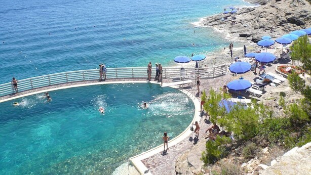 A clear blue swimming pool surrounded by a stone deck, with a rocky coastline and beach umbrellas in the background. - Four-room apartment in the Capo D'Arco area with direct access to the sea (Rio Marina)