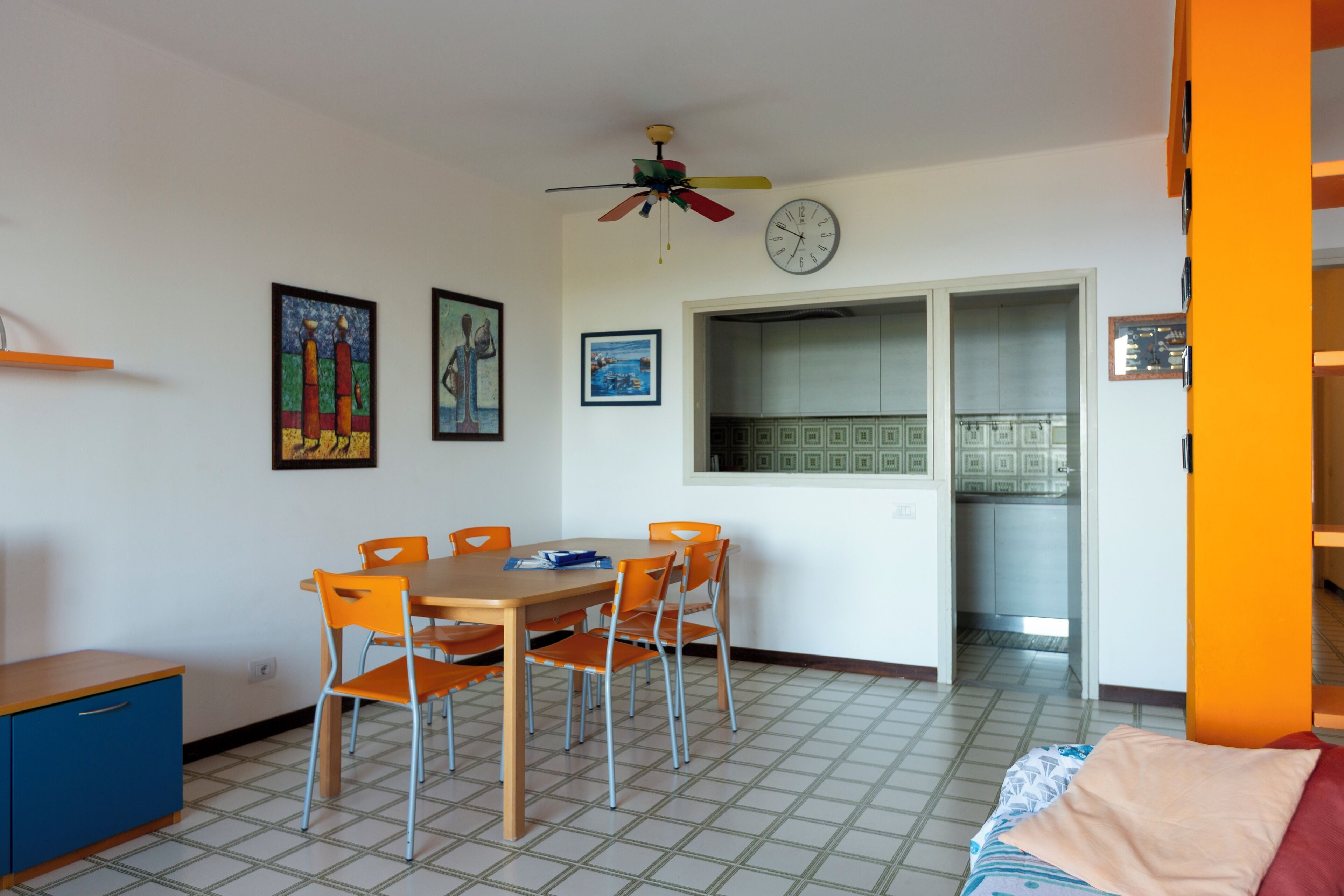 A dining area with a wooden table and orange chairs, a ceiling fan, and a clock on the wall.