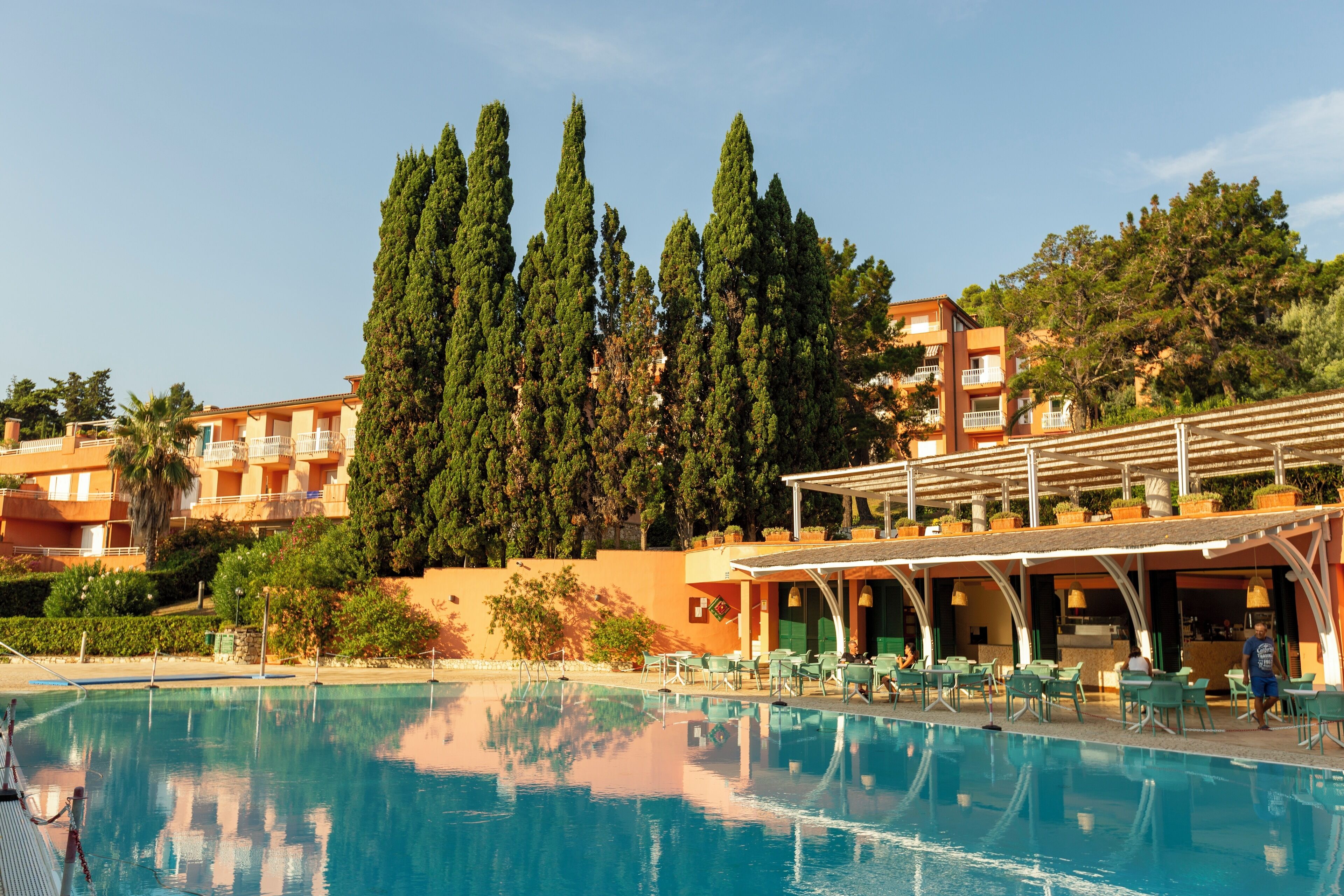 A swimming pool with lounge chairs and umbrellas, surrounded by tall trees and a building with balconies.