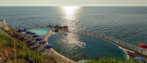 A coastal swimming pool with multiple sun loungers and umbrellas, surrounded by a railing and overlooking the sea.