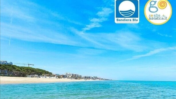 Une plage avec une eau turquoise et limpide, un rivage sablonneux et des bâtiments en arrière-plan.