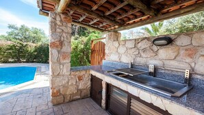 An outdoor kitchen with a stone wall, a built-in grill, and a view of a swimming pool. - Spacious Villa, Large Private Pool, Outdoor Jacuzzi, Attractive Gardens, 10 min walk to the Beach! (Latchi)