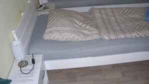 Una habitación con dos camas individuales, cada una con un colchón gris y una manta tejida. Hay una mesita de noche con una lámpara y un libro.