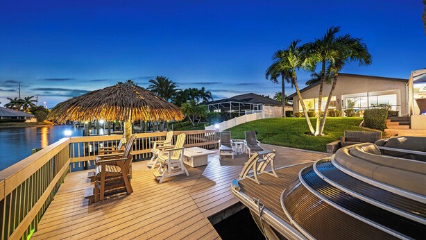 Une terrasse en bois avec des sièges d’extérieur, un parasol en chaume et un espace aménagé au bord de la piscine, agrémenté de palmiers.