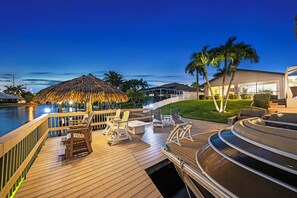 Una terraza de madera con mobiliario exterior, un toldo de paja y una zona junto a la piscina con palmeras.
