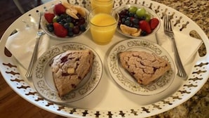 Een ontbijtplateau met twee scones, een schaal fruit en twee glazen sinaasappelsap op een bord met dessin.
