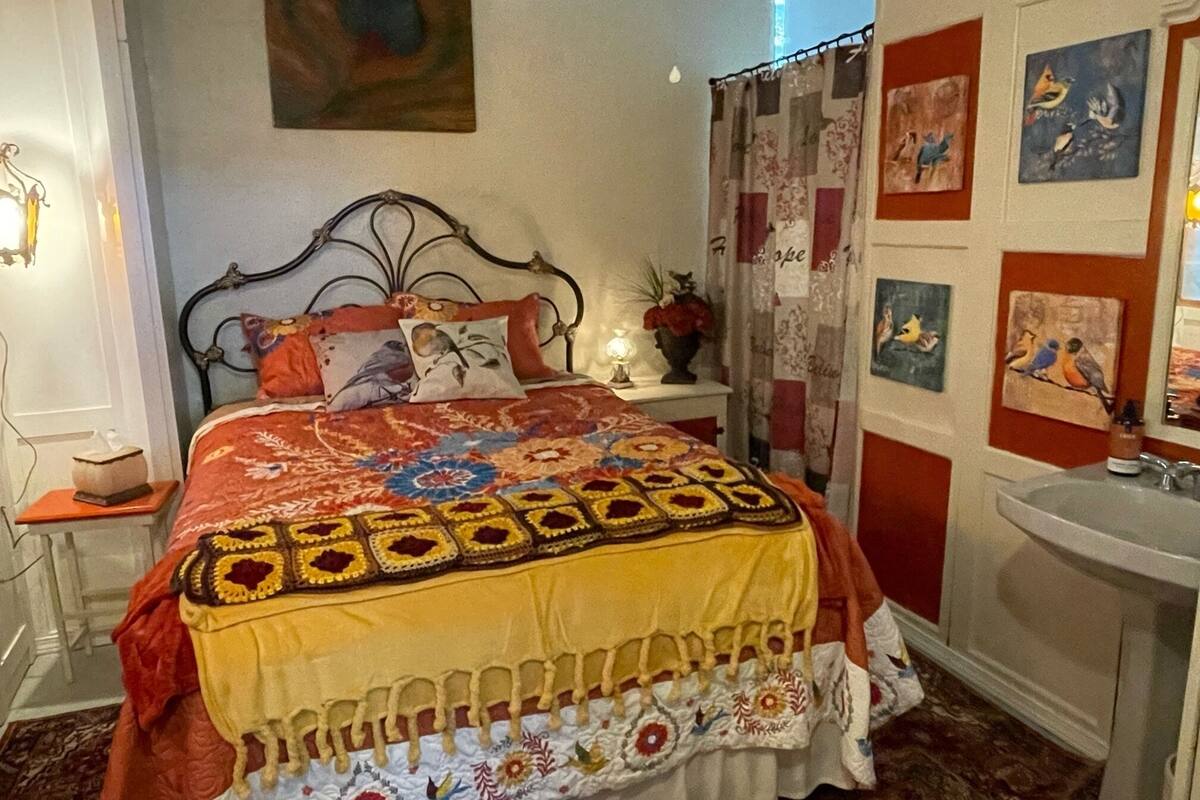 A bedroom with a bed, a nightstand, a lamp, a wall with artwork, and a bathroom with a sink.
