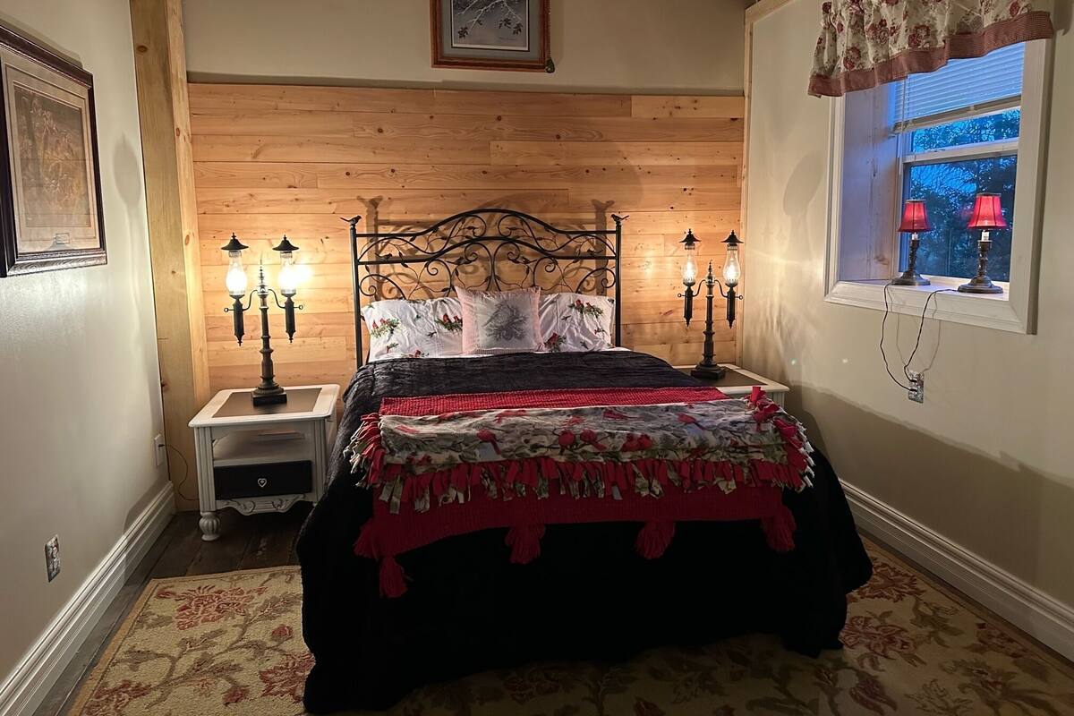 A bedroom with a wooden headboard, a bed with a floral bedspread, two bedside lamps, a nightstand, and a window with a view of trees.