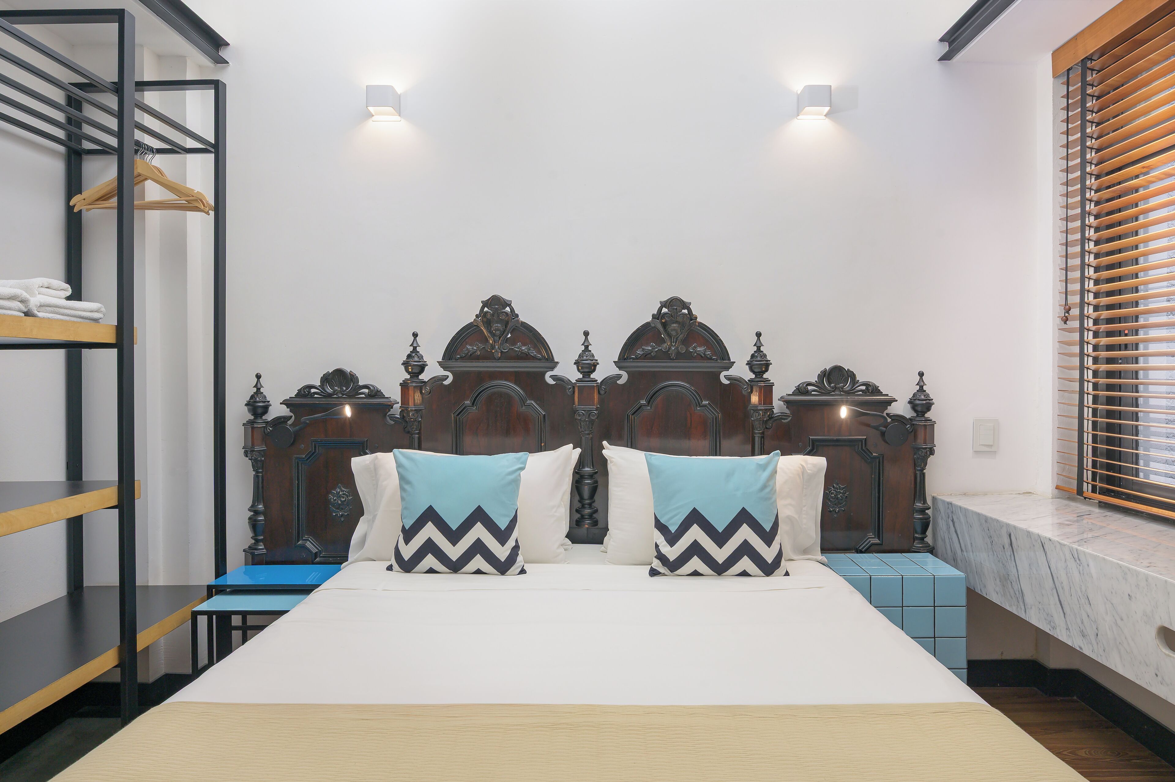 Una cama con cabecera de madera, dos almohadas, una mesita de noche y un estante con toallas dobladas.