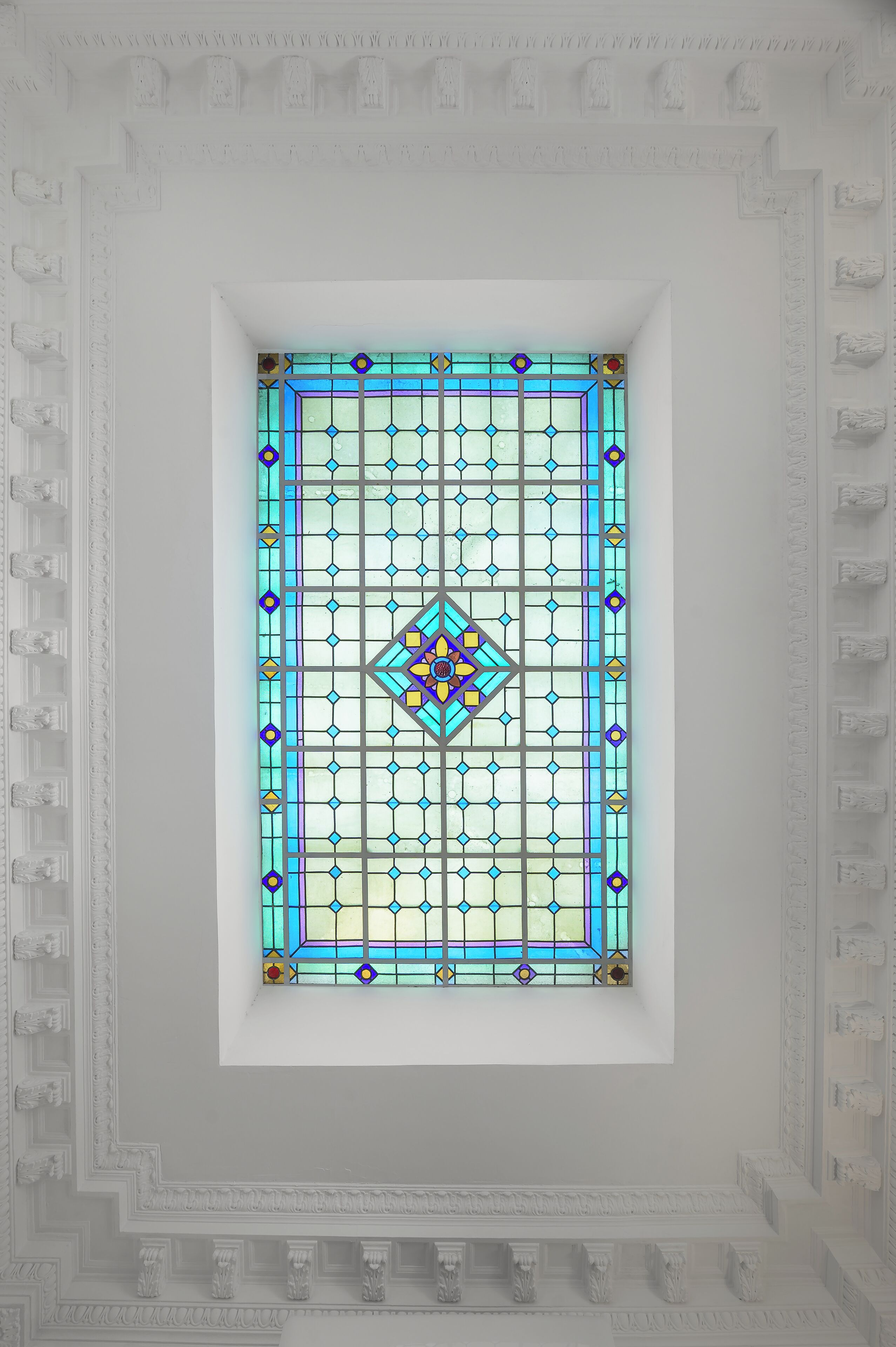 A stained glass window with geometric patterns and a central diamond design, set within a white-framed niche in a ceiling with decorative molding.