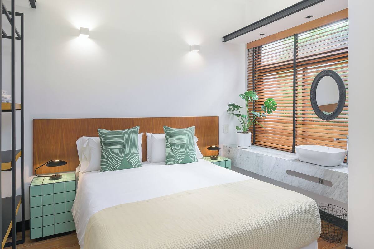 Un dormitorio moderno con una cama grande, una mesita de noche, un espejo y una ventana con persianas.