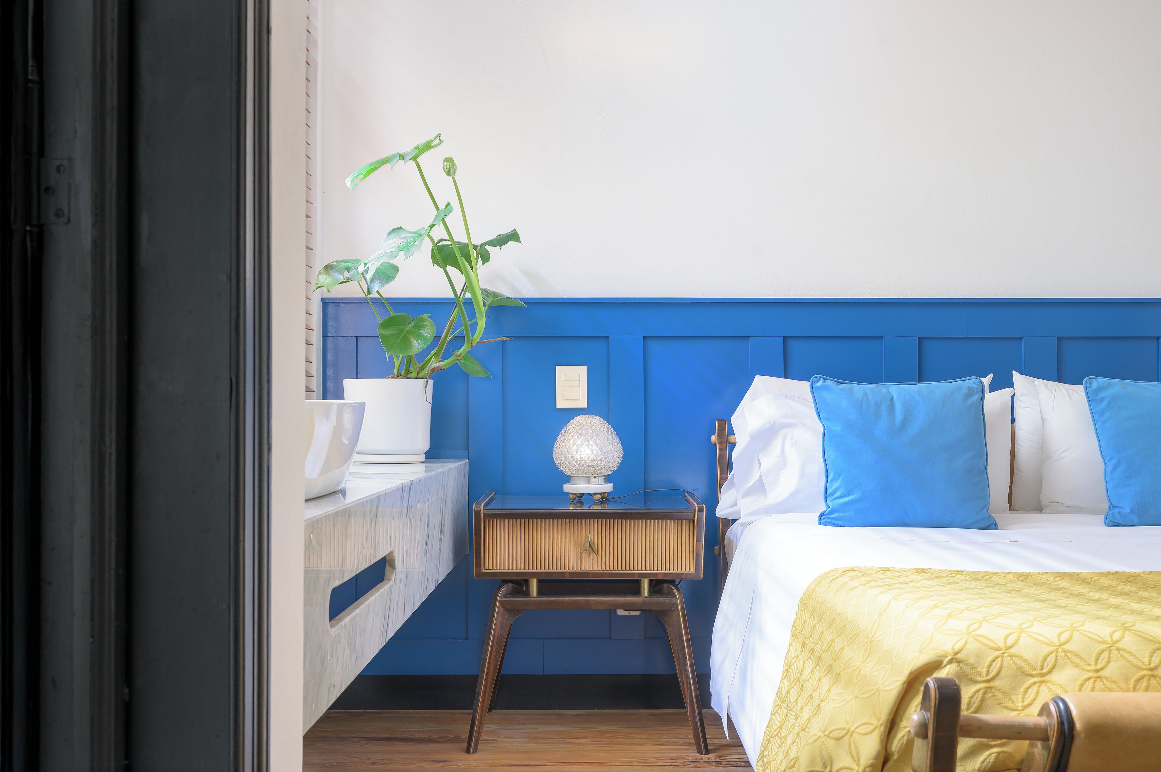 Un dormitorio con una cama, una mesita de noche con lámpara, una planta en maceta y un cabecero azul.