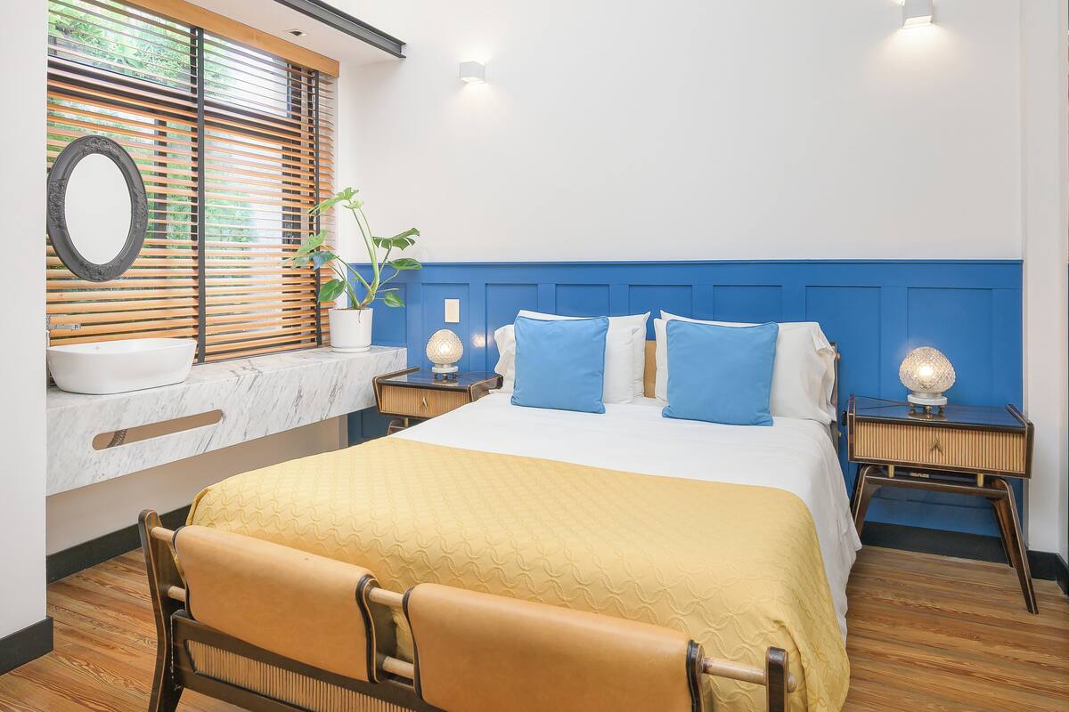 Un dormitorio moderno con una cama grande, una mesita de noche con lámpara, un espejo y una ventana con persianas.