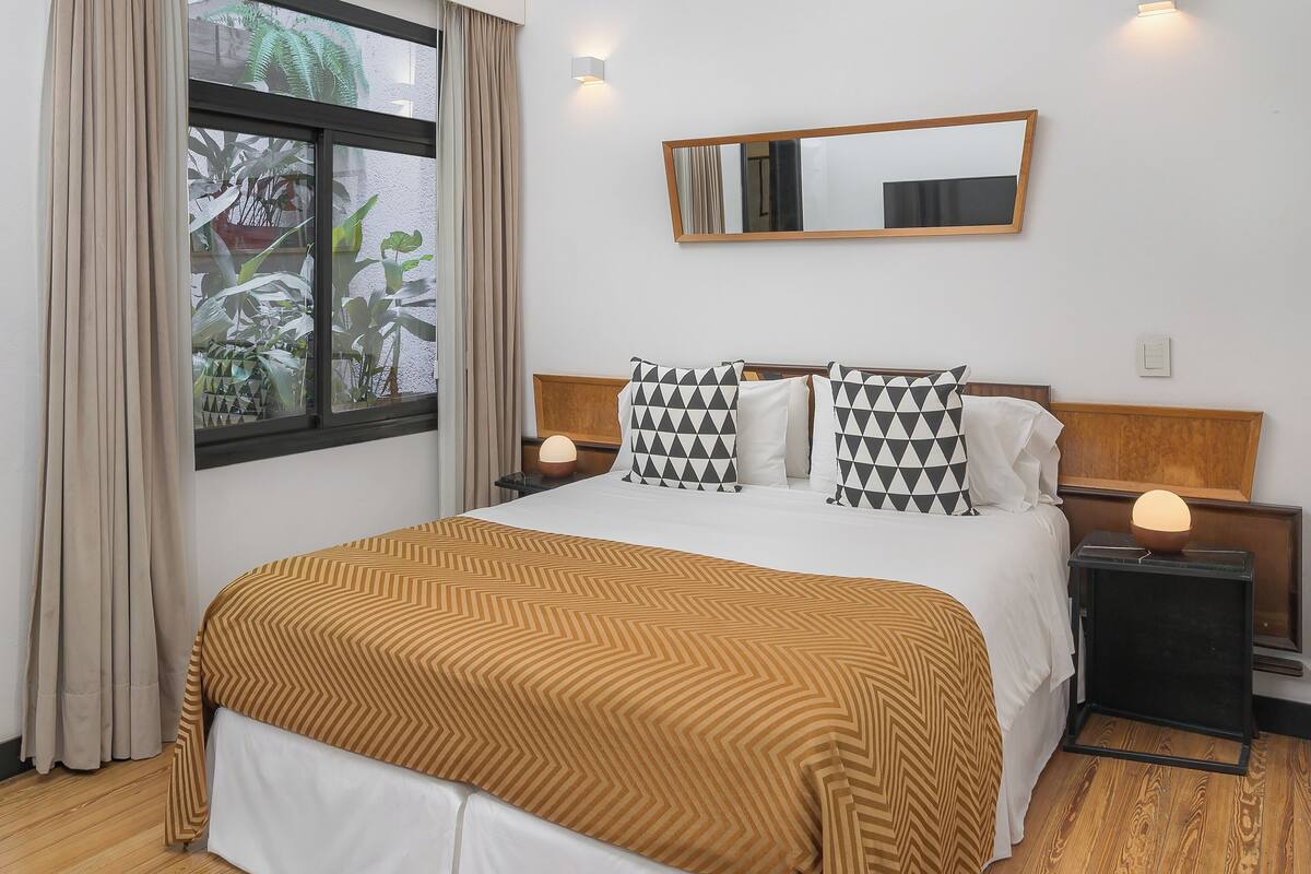 Una habitación de hotel con una cama grande, suelo de madera, una ventana con cortinas, una mesita de noche con una lámpara y un espejo instalado en la pared.