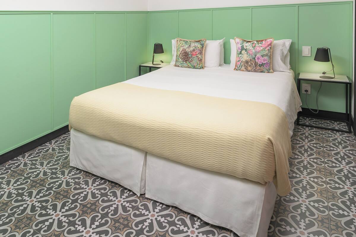 Habitación de hotel con una cama grande, dos mesitas de noche, pared verde, baldosas de suelo estampadas y una lámpara en el techo.