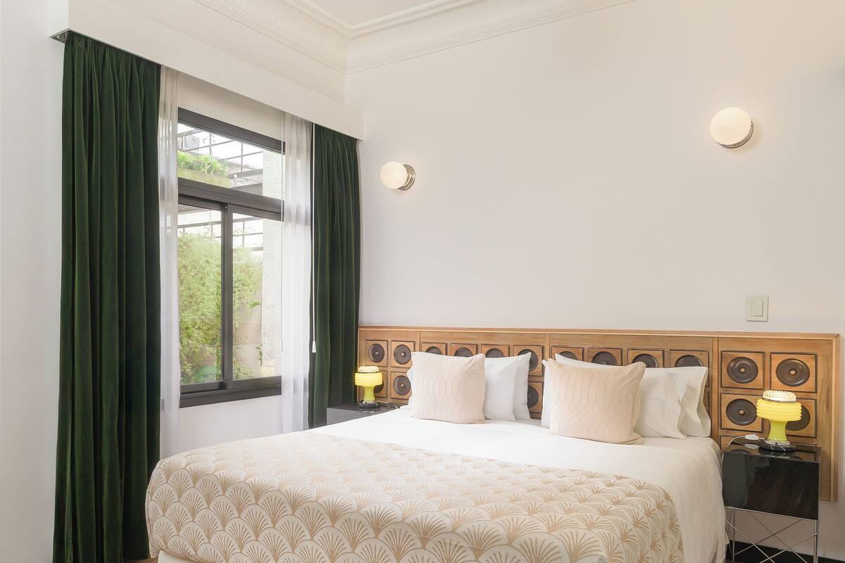 Habitación de hotel con una cama grande, cortinas verdes, cabecera de madera, dos lámparas de noche y vistas por la ventana.