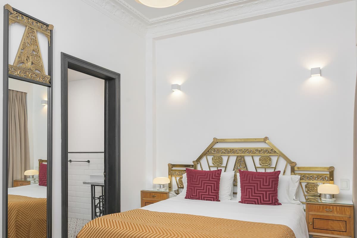 Un dormitorio con una cama grande, suelo de madera, un espejo, una mesita de noche e iluminación en la pared.