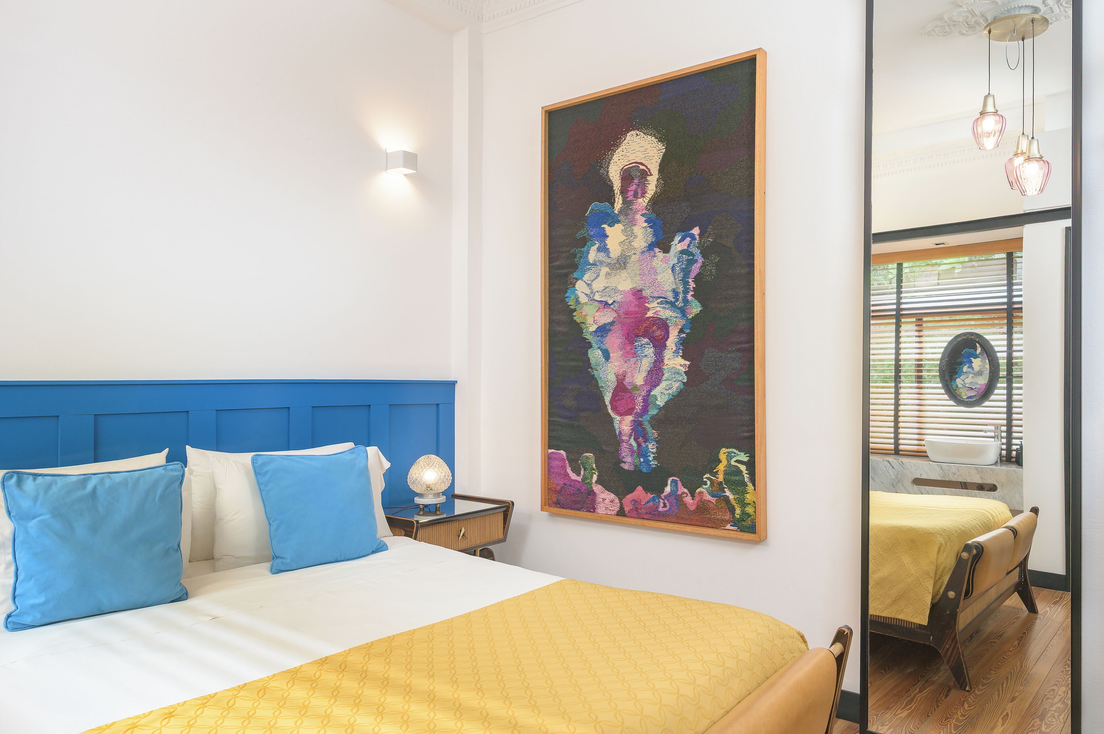 A bedroom with a bed, blue and white pillows, a yellow blanket, a painting, and a window with blinds.