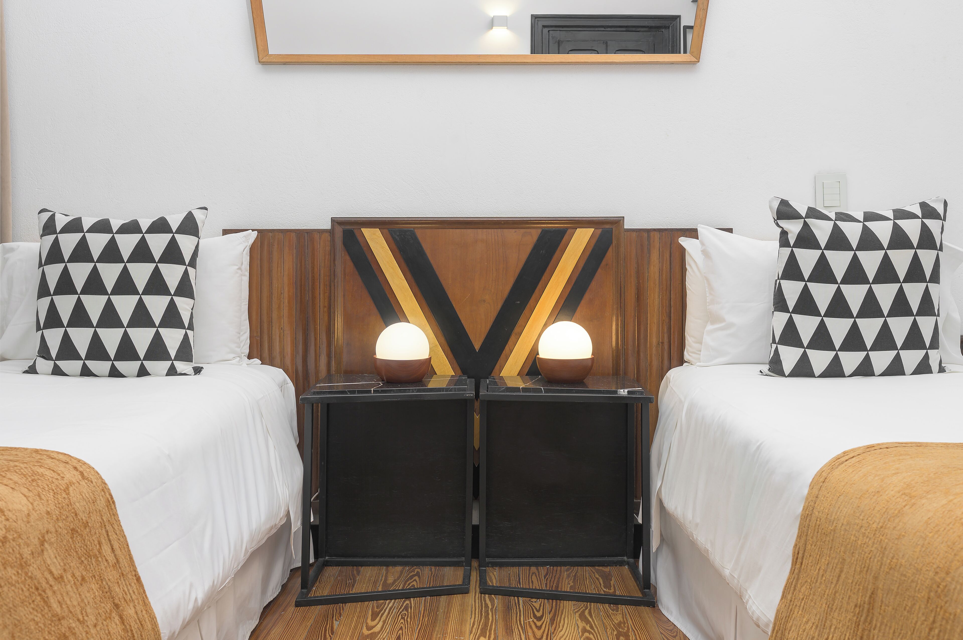 Quarto de hotel com duas camas, cada uma com almofada estampada em preto e branco, cabeceira de madeira e uma pequena mesa com duas luminárias entre as camas.