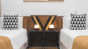 Quarto de hotel com duas camas, cada uma com almofada estampada em preto e branco, cabeceira de madeira e uma pequena mesa com duas luminárias entre as camas.