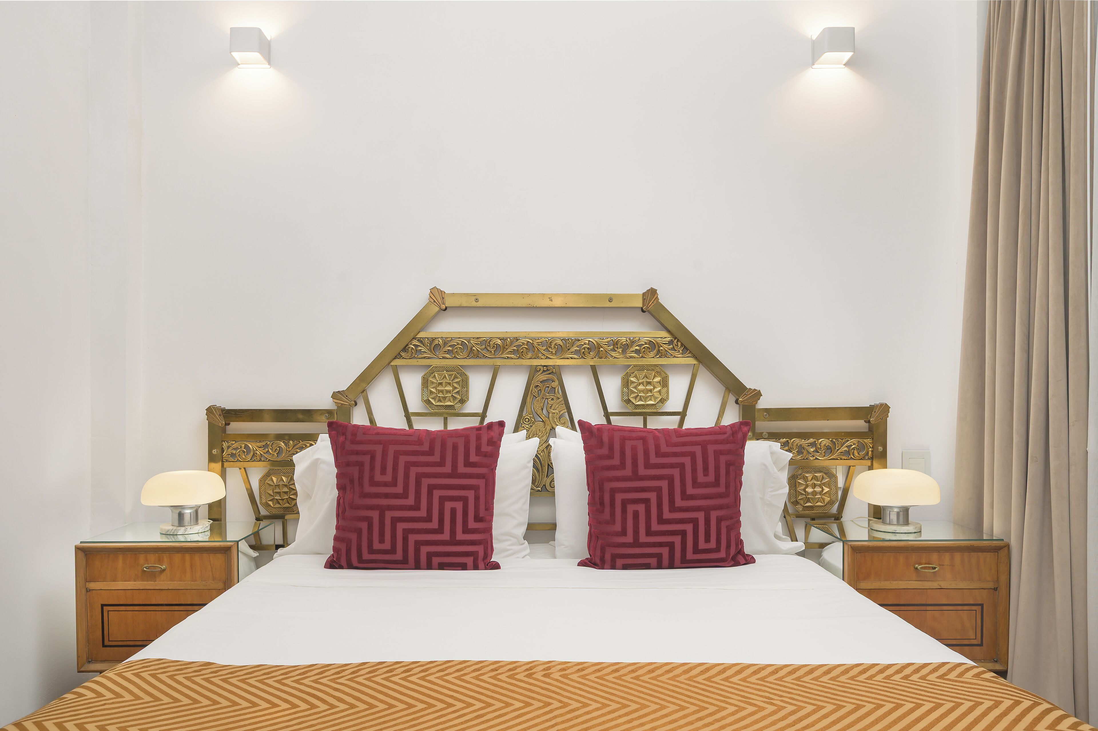 Una cama bien hecha con una colcha estampada, dos almohadones decorativos, un cabecero de madera con intrincados grabados y lámparas de noche sobre mesitas de madera.