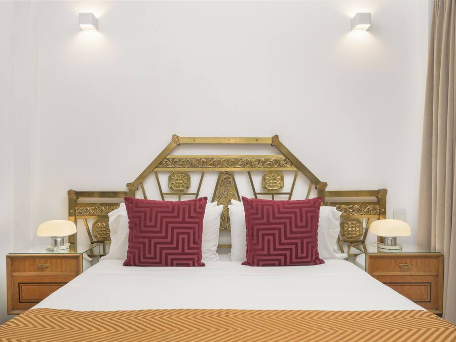 Una cama bien hecha con una colcha estampada, dos almohadones decorativos, un cabecero de madera con intrincados grabados y lámparas de noche sobre mesitas de madera.