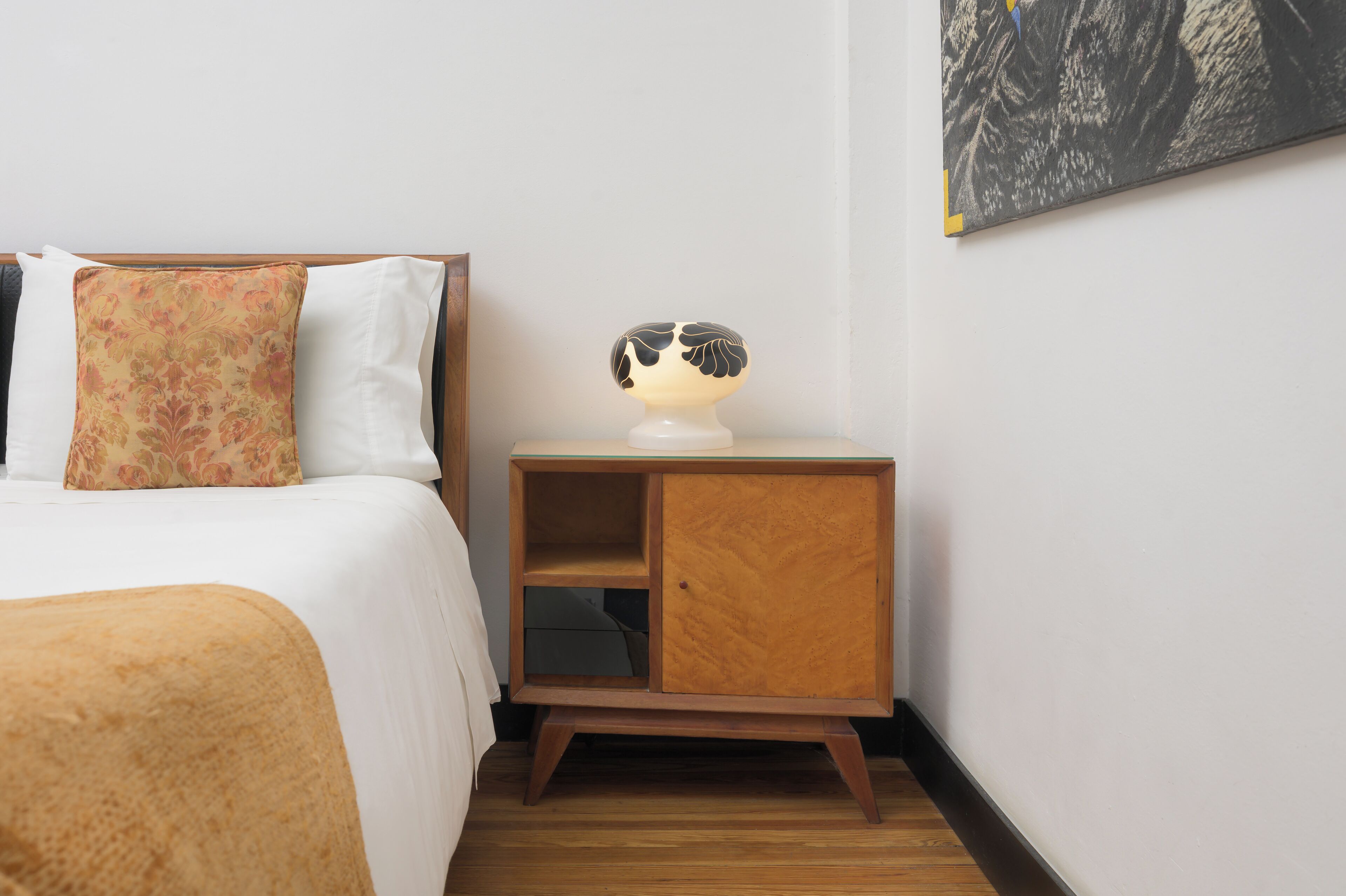 Una cama bien tendida con un cojín floral y una mesita de noche con una lámpara y un pequeño armario de madera.
