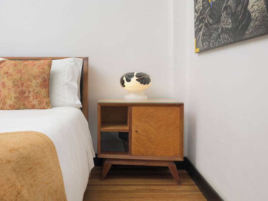 Una cama bien tendida con un cojín floral y una mesita de noche con una lámpara y un pequeño armario de madera.