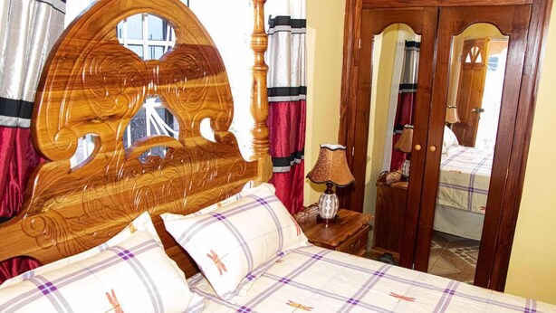 A bedroom with a wooden headboard, a bed with plaid bedding, a bedside lamp, and a mirror.