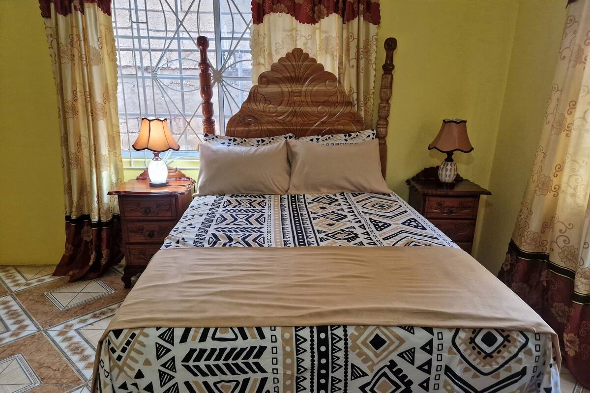A bed with a patterned bedspread, two bedside tables with lamps, curtains, and a window with a metal grid.