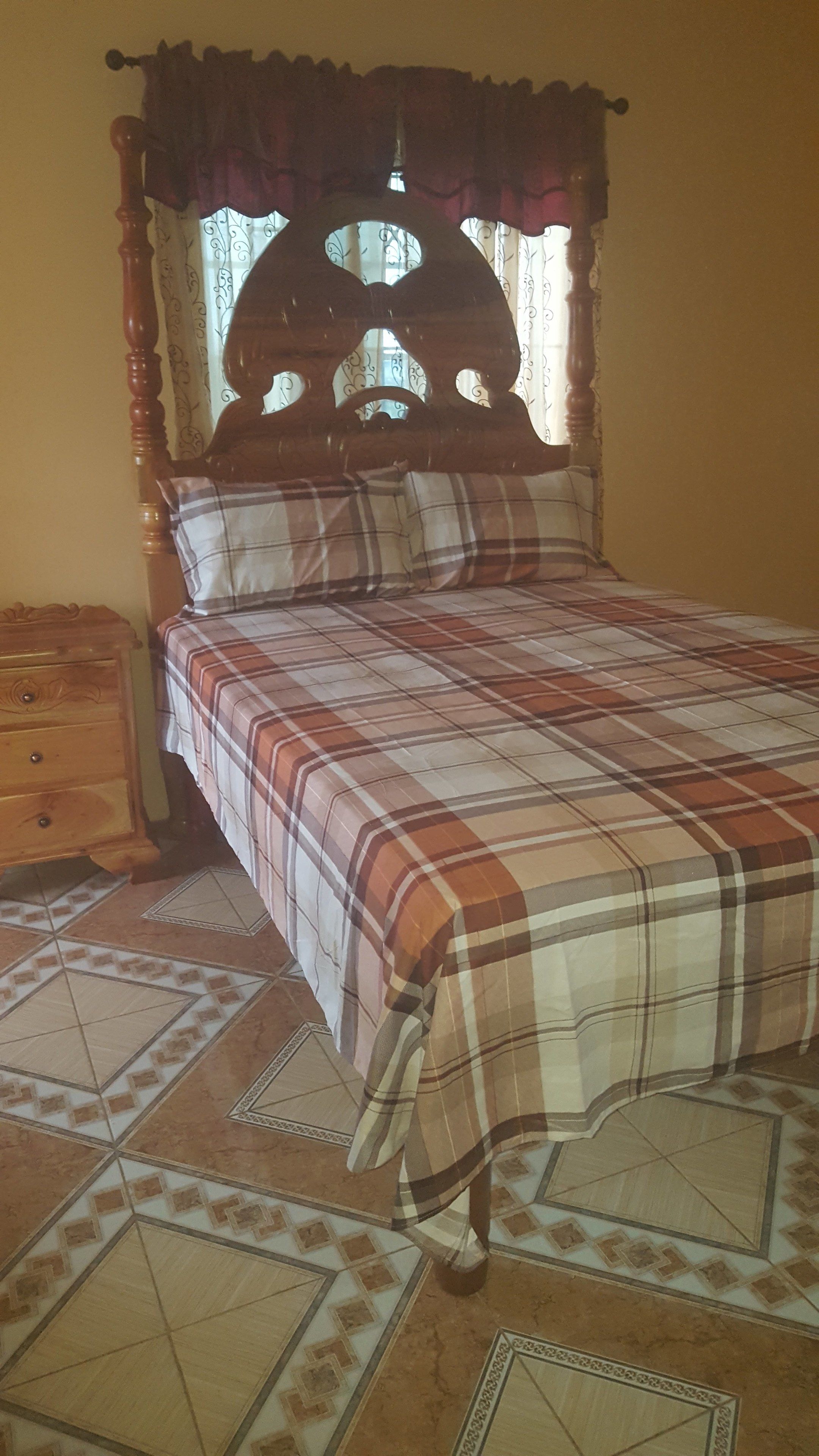 Una cama con colcha de cuadros, cabecera de madera y mesita de noche con cajón.
