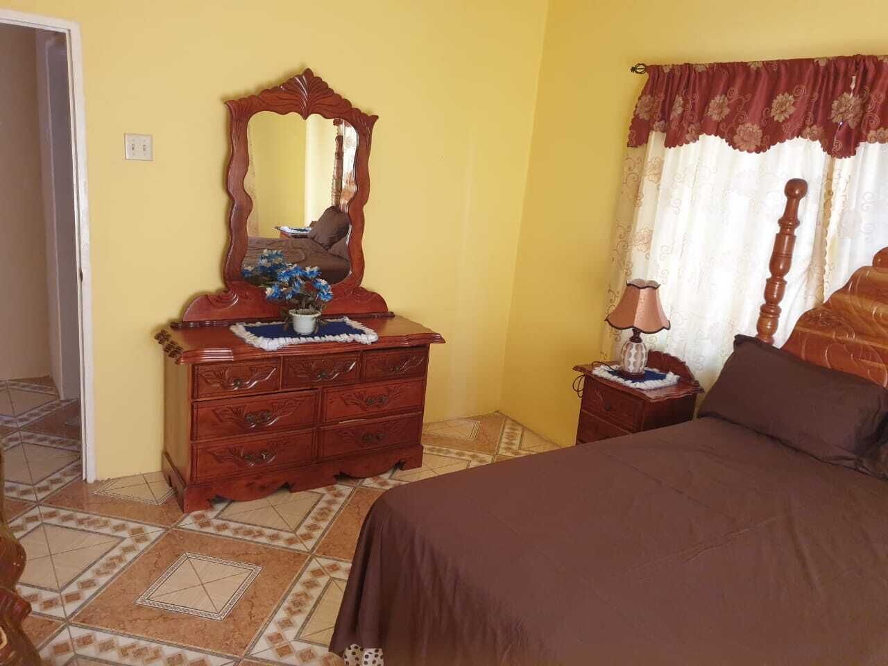 A bedroom with a wooden dresser, a bed with a brown bedspread, a nightstand with a lamp, and a window with curtains.